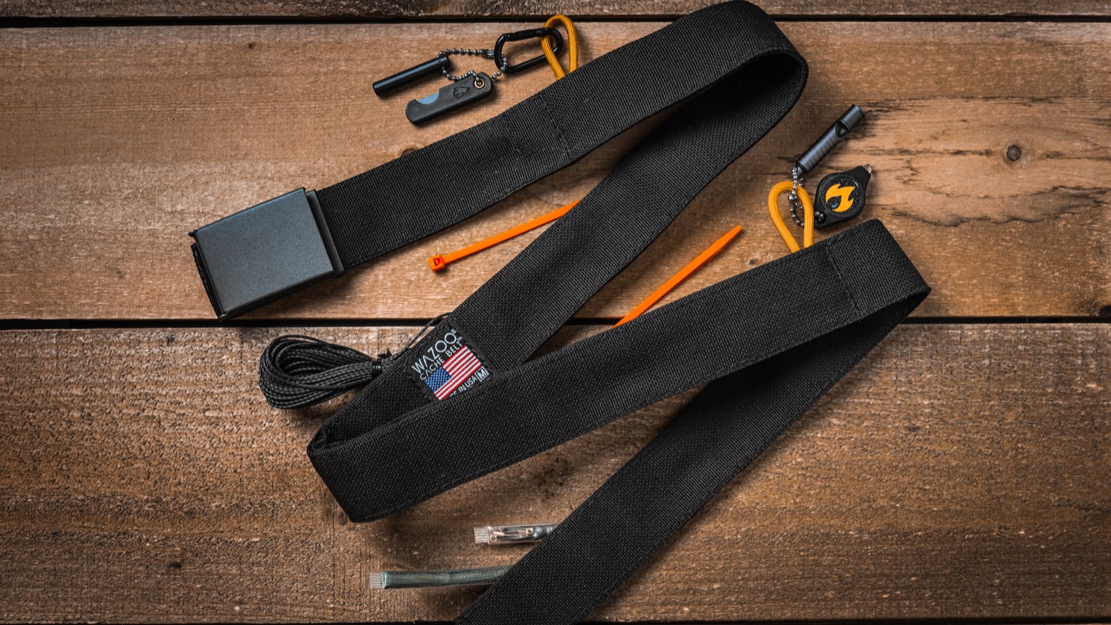 Wazoo Cache Belt laid out on a wood table with the survival kit that fits in its hidden pocket: ferro rod, whistle, light, cord and zip ties